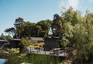 Un lodge con terraza de madera, rodeado de vegetación y árboles, situado junto a un lago tranquilo.