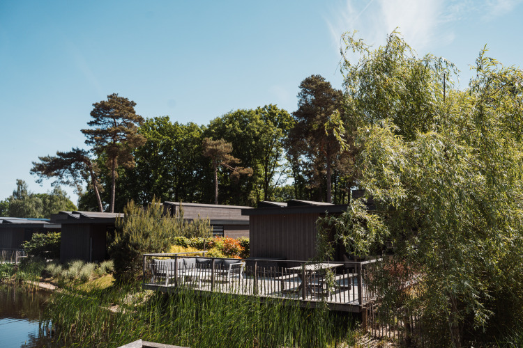 A lodge with a wooden deck, surrounded by lush greenery and trees, located beside a tranquil lake.