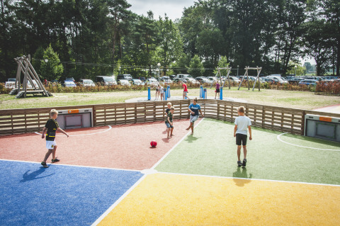 Niños juegan fútbol en una cancha colorida en Familievakantiepark Krieghuusbelten con área de juegos y autos al fondo.