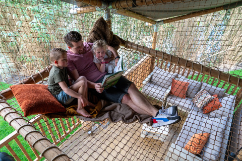 Vader leest een boek aan twee kinderen in een net-hangmat in een safaritent op Krieghuusbelten, Nederland.