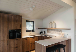 Cocina moderna de glamping con armarios de madera, encimeras blancas y dos taburetes negros.