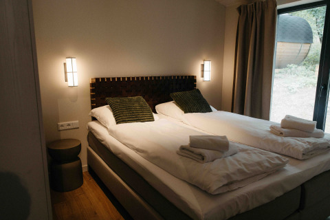 Glampingverblijf met twee aparte bedden, groene kussens, propere handdoeken en groot raam met uitzicht.