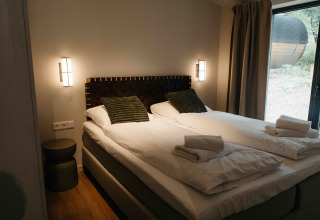 Glampingverblijf met twee aparte bedden, groene kussens, propere handdoeken en groot raam met uitzicht.