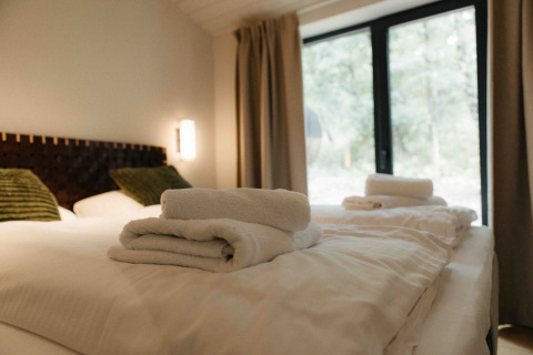 Glamping verblijf met knus bed, verse handdoeken, groene kussens en een groot raam met zicht op de natuur.
