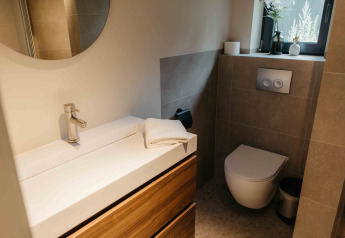 Baño moderno en alojamiento glamping con inodoro de pared, lavabo, gran espejo y ventana luminosa.