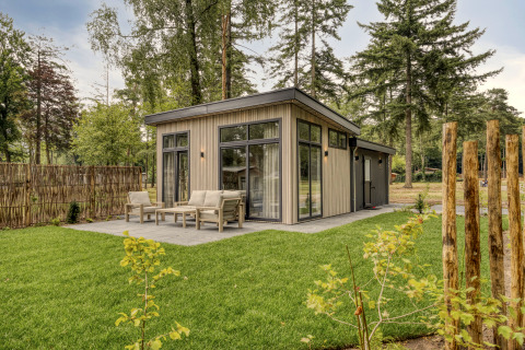Moderne lodge met grote ramen en terras, omgeven door een groene grasmat en hoge bomen.