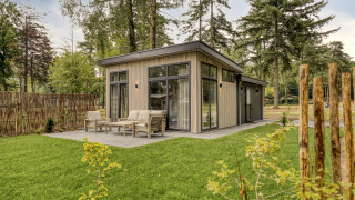 Lodge moderne avec de grandes fenêtres et terrasse aménagée, entourée d’herbe verte et de grands arbres.