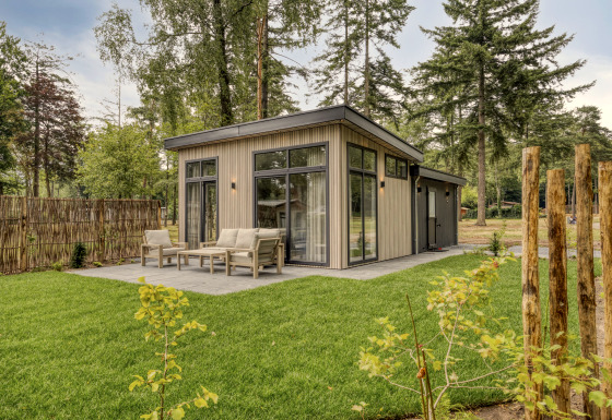 Moderne lodge met grote ramen en terras, omgeven door een groene grasmat en hoge bomen.