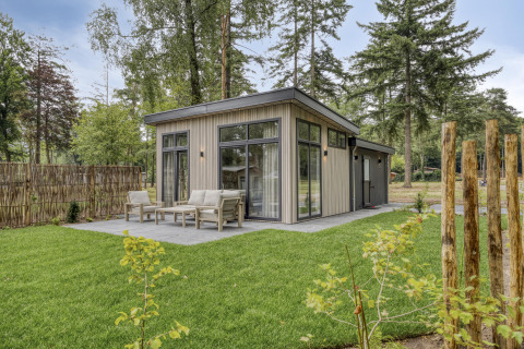 Modern lodge with large windows and patio seating, surrounded by lush green lawn and tall trees.