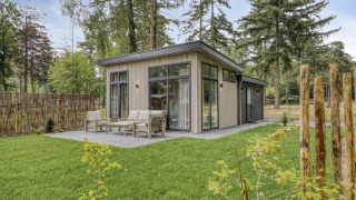 Moderne lodge met grote ramen en terras, omgeven door een groene grasmat en hoge bomen.
