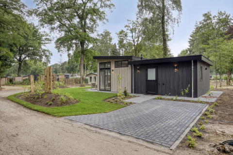 Lodge moderne à façade sombre et grandes baies vitrées, entouré d’arbres et d’une pelouse soignée.