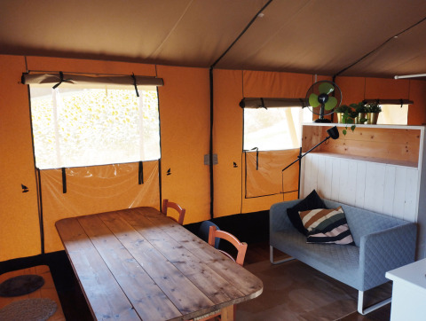 Interieur van een safaritent met houten tafel, bank, planten en ramen op Camping Suasa in Italië.