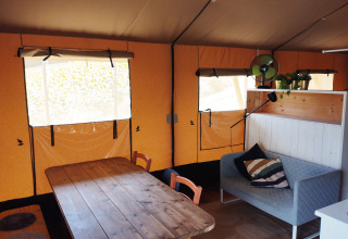 Interieur van een safaritent met houten tafel, bank, planten en ramen op Camping Suasa in Italië.