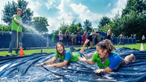 Zwei junge Frauen rutschen lachend auf einer Wasserrutsche im Freien, während ein Mann sie abspritzt.