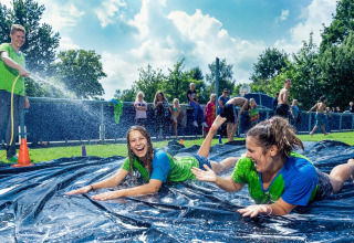 Zwei junge Frauen rutschen lachend auf einer Wasserrutsche im Freien, während ein Mann sie abspritzt.