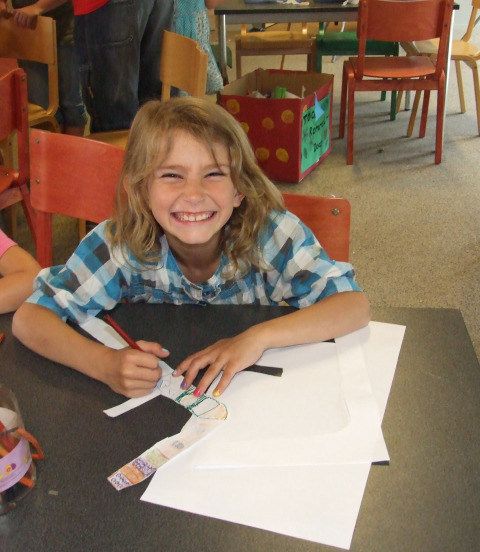 A smiling child with long hair draws with colored pencils at a table in a cheerful classroom setting.