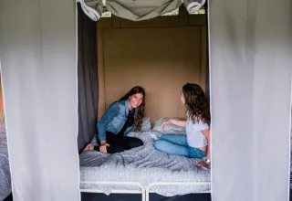 Deux jeunes femmes assises et souriantes sur un lit sous une tente au parc de vacances ’t Strandheem, Groningen.