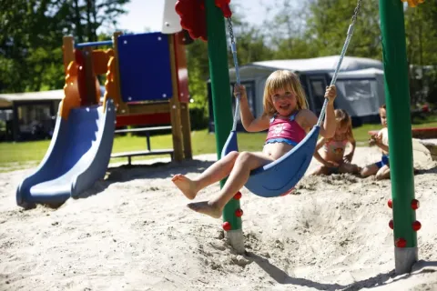 Kinderen spelen op een zandige speeltuin met schommels en glijbaan bij vakantiepark ’t Strandheem.