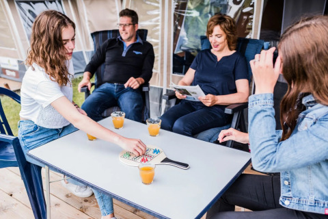 Een gezin geniet in een safaritent, speelt een bordspel en drinkt samen sap rond een buitentafel.