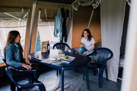 Due donne si rilassano in una tenda safari, sedute a un tavolo con vino, riviste e sedie di plastica.