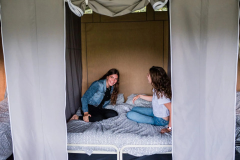 Twee vrouwen zitten pratend op een bed in een safaritent met open gordijnen en een ontspannen sfeer.