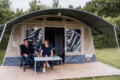 Twee mensen ontspannen met drankjes op een houten veranda voor een grote safaritent met groene omringing.