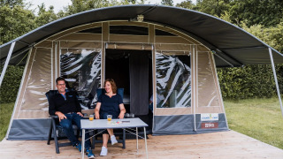 Zwei Personen sitzen entspannt mit Getränken vor einem großen Safarizelt auf einer Holzterrasse.