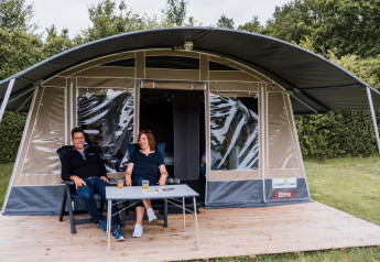 Two people relax with drinks on a wooden deck in front of a large safari tent surrounded by greenery.