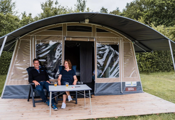 Due persone si rilassano con delle bevande su una pedana di legno davanti a una grande tenda safari.