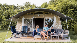 Una famiglia si rilassa su sedie davanti a una tenda safari moderna circondata da alberi e prato verde.
