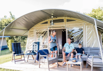 Gezin ontspant buiten aan een safaritent met tuinmeubels, drankjes en zonlicht in de natuur.