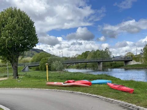 Kayaks on grass by the river with a bridge and scenic cloudy sky at Camping Gritt, Diekirch, Luxembourg.