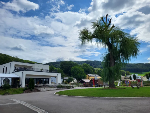 Camping Gritt a Diekirch, Lussemburgo, con edifici moderni, grande albero e parco giochi in una giornata serena.
