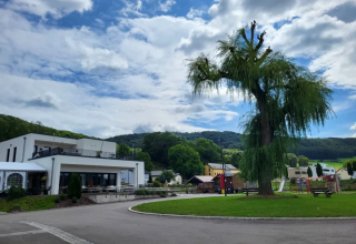 Camping Gritt i Diekirch, Luxembourg, med moderne bygninger, store træer og legeområde under blå himmel.