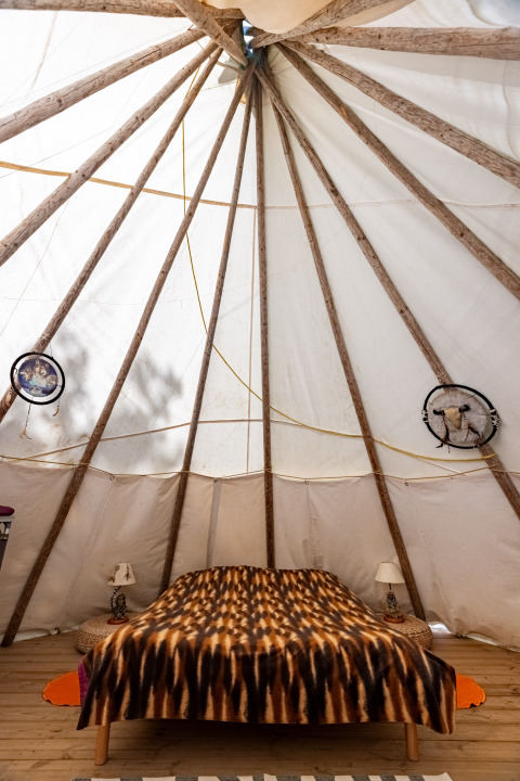 Vista interior de una tienda tipi con estructura de madera, cama doble y lámparas en Camping Des Randonneurs en Francia.