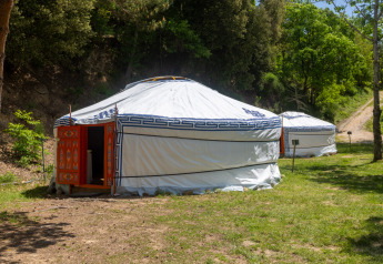 Twee traditionele yurts met witte stof en blauwe motieven staan op een groene, bosrijke camping overdag.