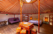 Interior of a traditional yurt with colorful painted furniture, a bed, cozy lighting and wooden flooring.