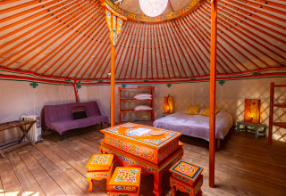 Interior of a traditional yurt with colorful painted furniture, a bed, cozy lighting and wooden flooring.