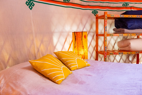 Interno accogliente di una yurta glamping con cuscini gialli, letto morbido e scaffali decorativi variopinti.