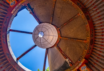 Looking up through the decorated roof opening of a traditional yurt with blue sky visible outside.