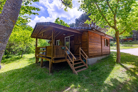 Wooden chalet with covered porch, surrounded by lush green trees and grass under a bright sky.