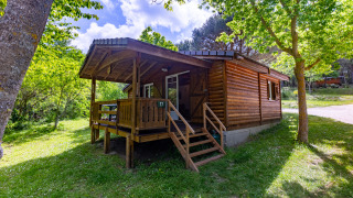 Wooden chalet with covered porch, surrounded by lush green trees and grass under a bright sky.