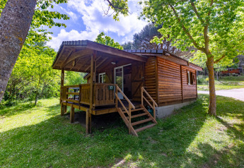 Wooden chalet with covered porch, surrounded by lush green trees and grass under a bright sky.