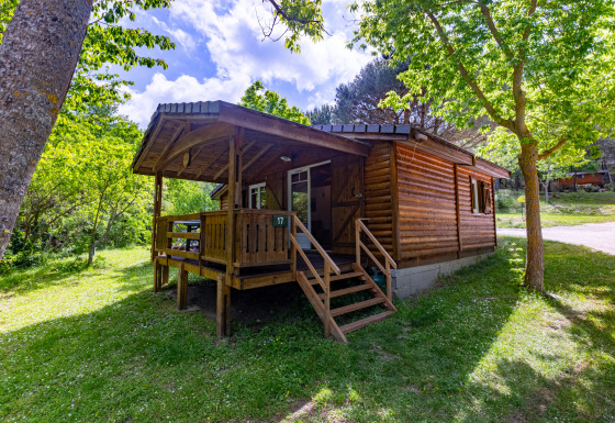 Wooden chalet with covered porch, surrounded by lush green trees and grass under a bright sky.