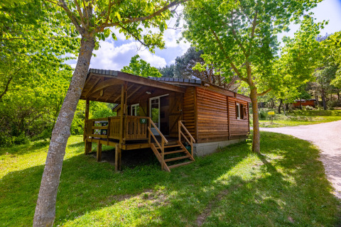 Wooden chalet with covered porch nestled among green trees at Camping Des Randonneurs in France in summer.