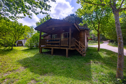 Wooden chalet at Camping Des Randonneurs in France, nestled among green trees and sunlit grassy area.