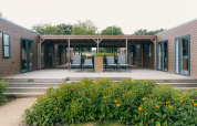 Udsigt til en moderne træhytte med terrasse, havestole og blomsterbede foran, omgivet af natur.