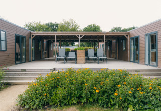 Blick auf eine moderne Holzhütte mit Terrasse, Liegestühlen und Blumenbeeten im Grünen.