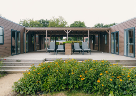 Uitzicht op een moderne lodge met houten terras, ligstoelen en een bloementuin aan de voorkant.