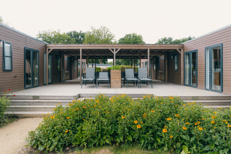 Blick auf eine moderne Holzhütte mit Terrasse, Liegestühlen und Blumenbeeten im Grünen.